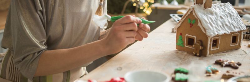 A person holding a piping bag of green frosting to decorate the mini gingerbread house sitting on the table surrounded by candy. 