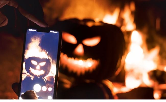 A lit up jack-o-lantern at night sitting in front of a campfire. A hand is holding a phone and taking a picture in front